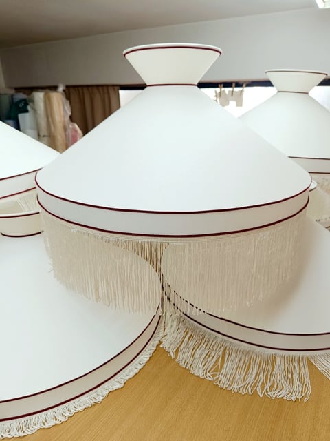 Multiple white dome-shaped lampshades with cream fringe trim and burgundy piping displayed on a wooden floor indoors